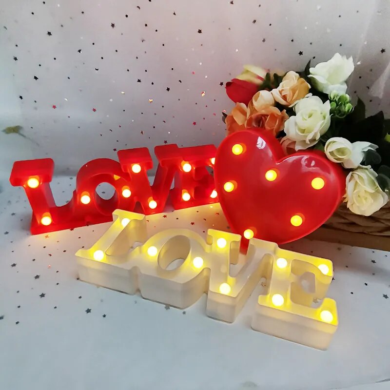 Two red nightlamps that read LOVE and a red heart lamp all illuminated by yellow LED's on a white tablecloth with silver glitter