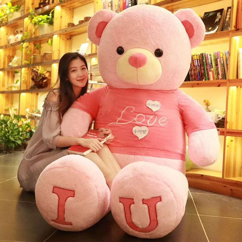 Large light pink teddy bear with dark pink shirt with I and U written on feet in dark pink lettering