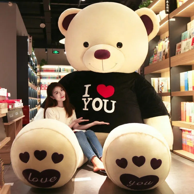 Large light beige teddy bear wearing black t-shirt reading I love you in white lettering with a red heart