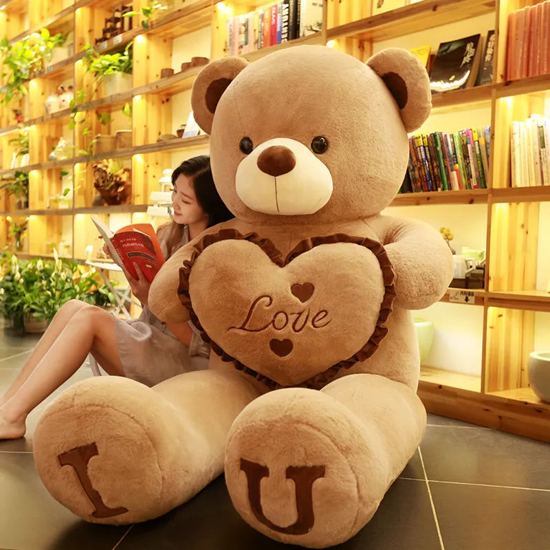 Large light brown teddy bear holding brown heart that reads love with I and U written on feet in brown lettering