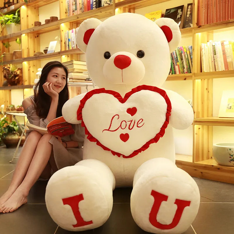 Large light beige teddy bear holding beige heart that reads love with I and U written on feet in red lettering