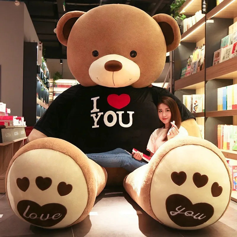 Large light brown teddy bear wearing black t-shirt reading I love you in white lettering with a red heart