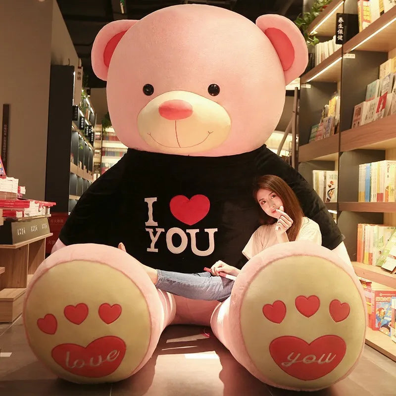 Large light pink teddy bear wearing black t-shirt reading I love you in white lettering with a red heart