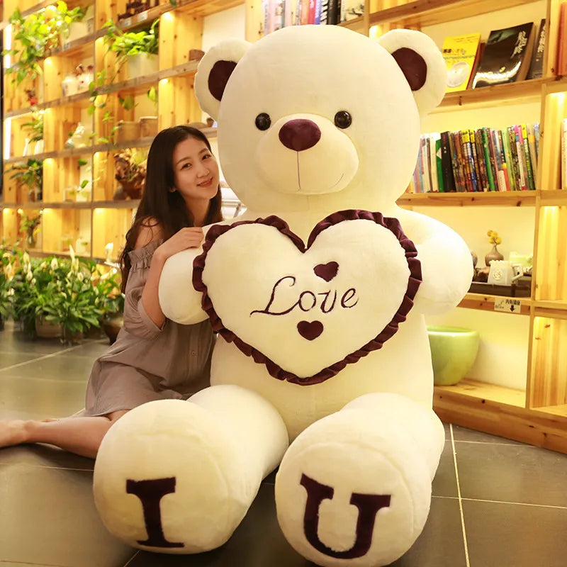 Large light beige teddy bear holding beige heart that reads love with I and U written on feet in brown lettering