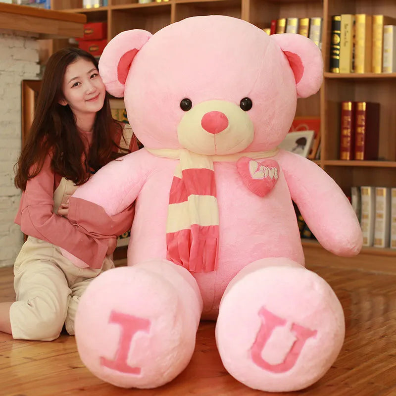 Large light pink teddy bear wearing brown dark pink scarf with I and U written on feet in pink lettering