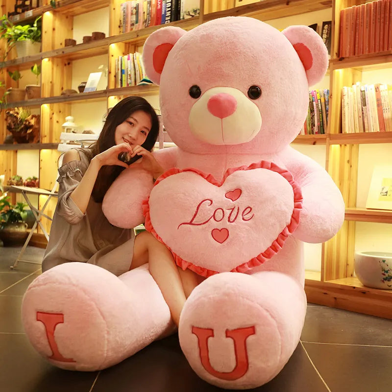 Large light pink teddy bear holding pink heart that reads love with I and U written on feet in pink lettering